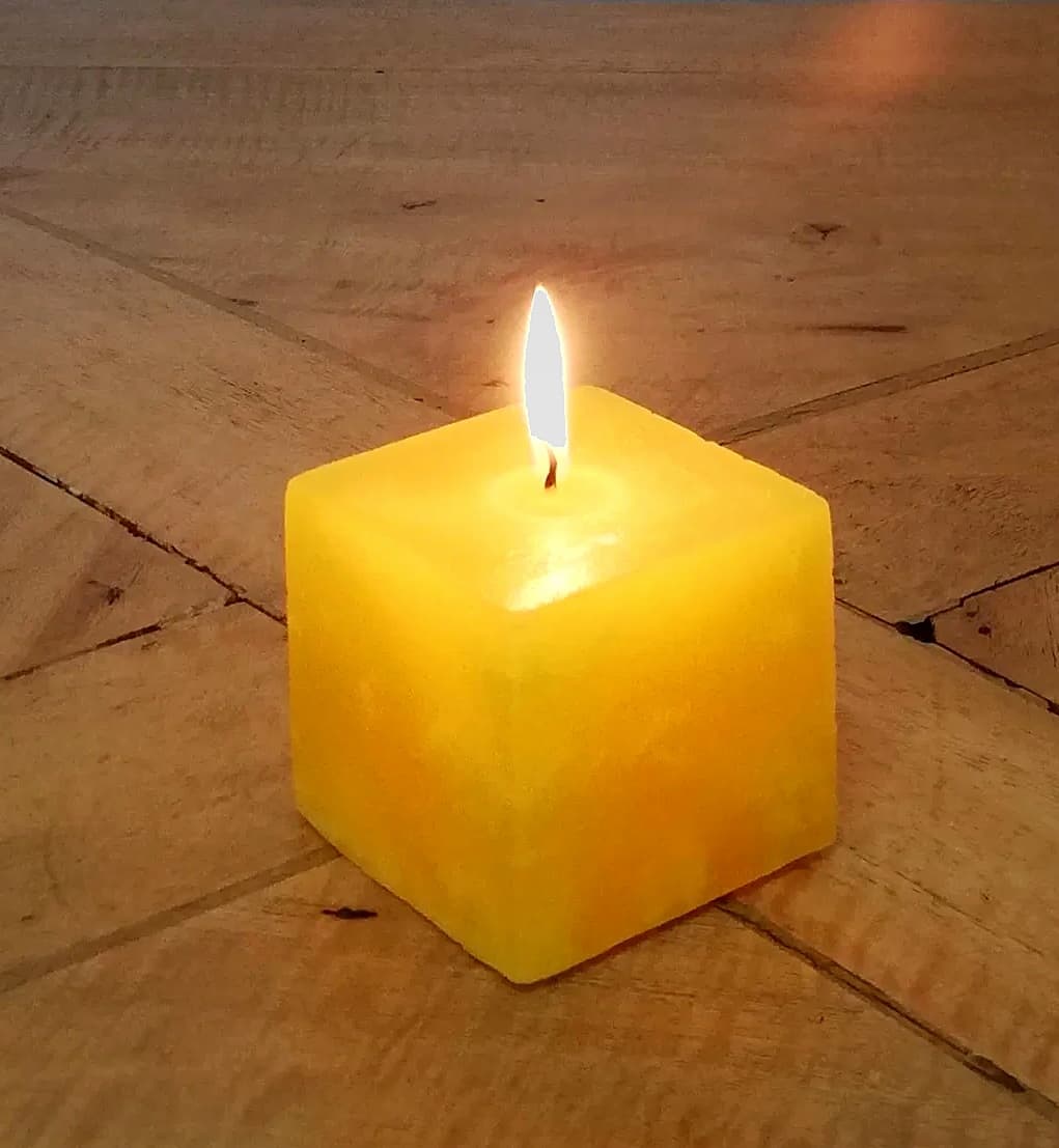 A lit square pillar candle in warm citrus-orange wax, standing on a wooden table.